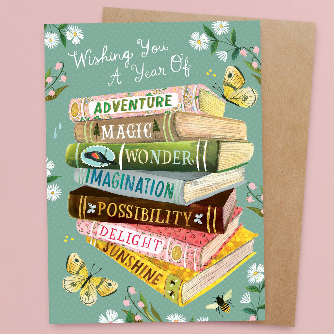 A greeting card with illustrated books stacked on each other, labeled Adventure, Magic, Wonder, Imagination, Possibility, Delight, Sunshine; surrounded by flowers, butterflies, and the text “Wishing You A Year Of.”.