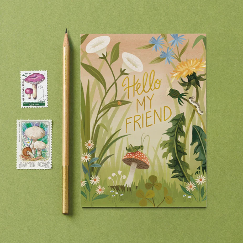 A Friendship Card by Carrie Shryock shows the phrase "Hello My Friend" amidst wildflowers, grass, a caterpillar, a ladybug, and a mushroom with a grasshopper on top. The card is set against a green background with a pencil and two flower postage stamps. 