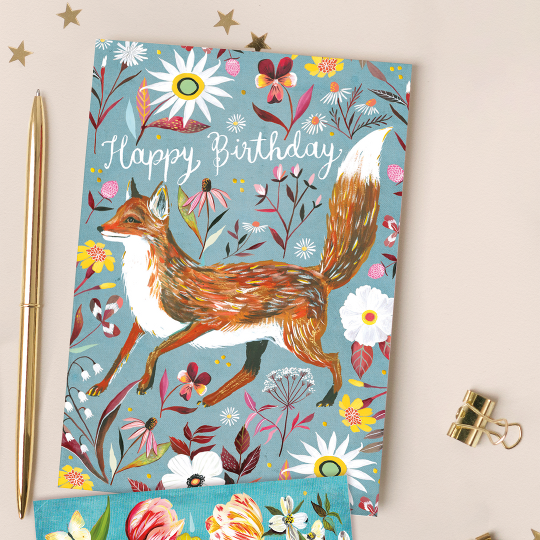 A beautiful birthday card showing a fox strolling through vibrant flowers and against a blue backdrop with the words "Happy Birthday" in white cursive with gold foil accents. The card sits on a cream background with a gold pen, paperclip, and stars. 