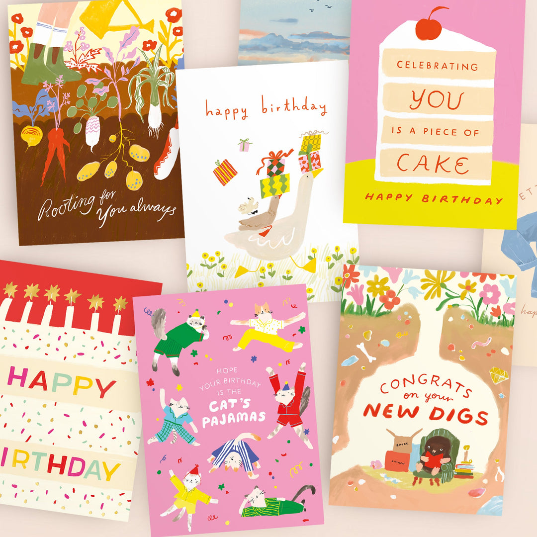 A woman in a floral dress sits at a table with tulips, greeting cards, art supplies, and a cup. She smiles at the camera; colorful cards and a landscape drawing decorate the wall behind her.