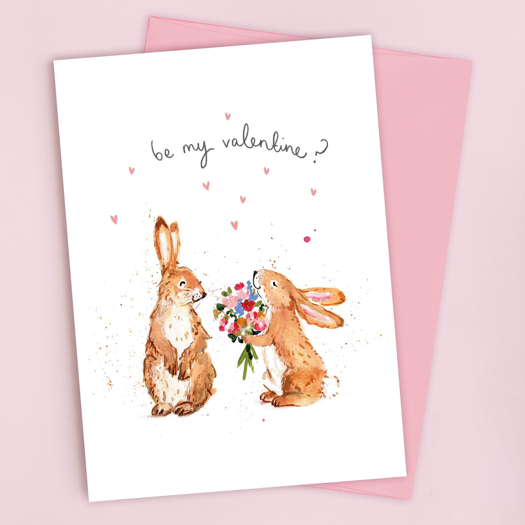 A card with two illustrated rabbits; one holds a bouquet of flowers, asking, be my valentine? Small pink hearts are scattered above them. A pink envelope lies underneath.